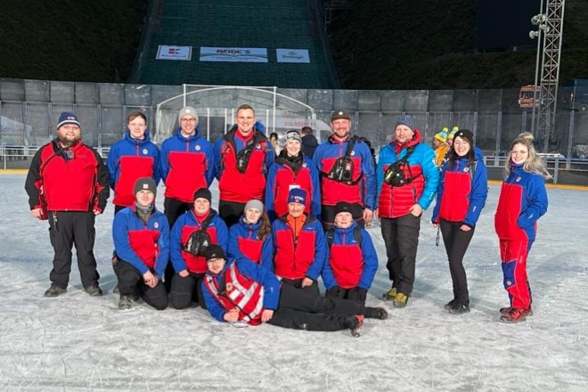 Kameradinnen und Kameraden der Bergwacht Klingenthal/Aschberg stehen auf der Eisfläche und tragen Ihre Dienstuniform. Gruppenbild am Abend unter Flutlicht.